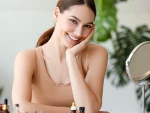 smiling woman sitting at table with bottles of essential oil in room