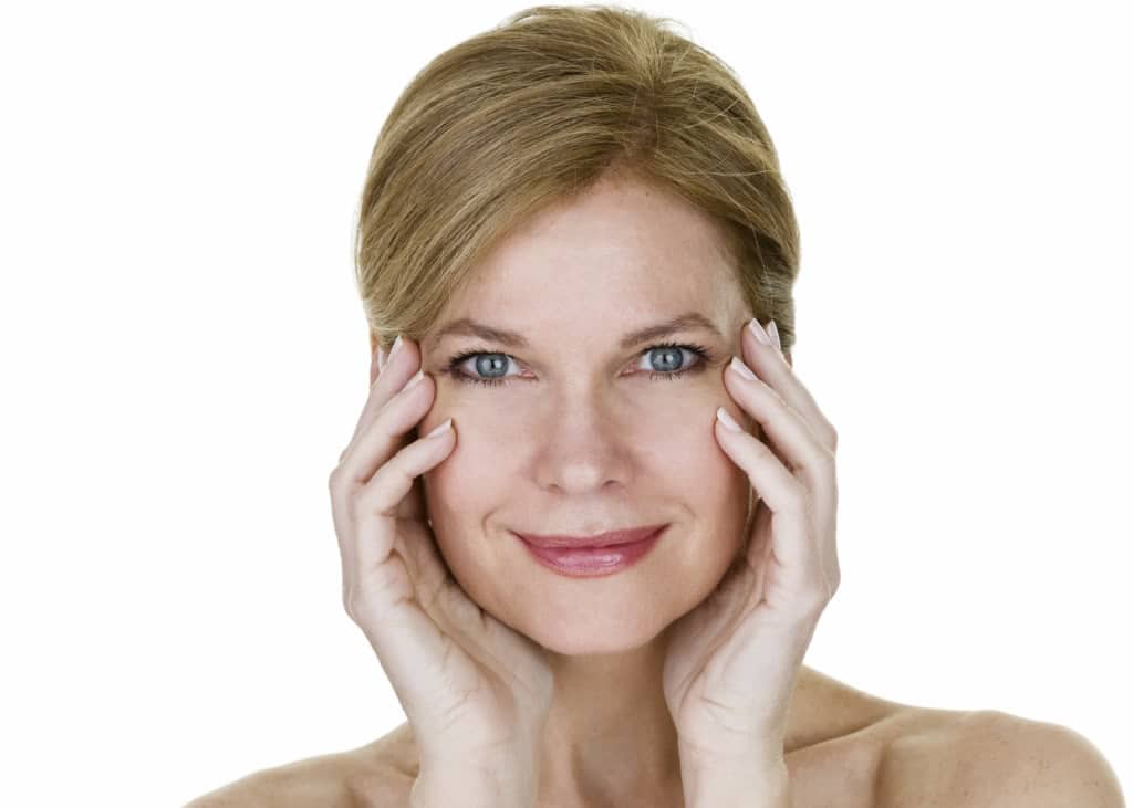 Middle-aged woman with glowing skin touching her face and smiling, showing smoother nasolabial lines from dermal filler treatment for smile lines.