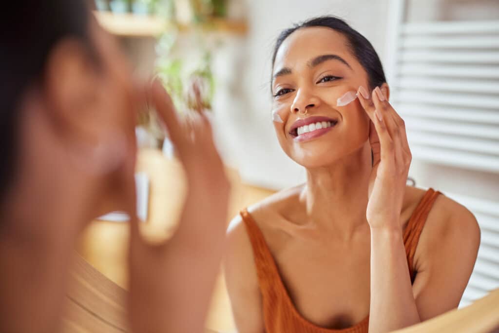 Woman applying facial skincare in a mirror as part of a sun-protection routine after a skin rejuvenation treatment.