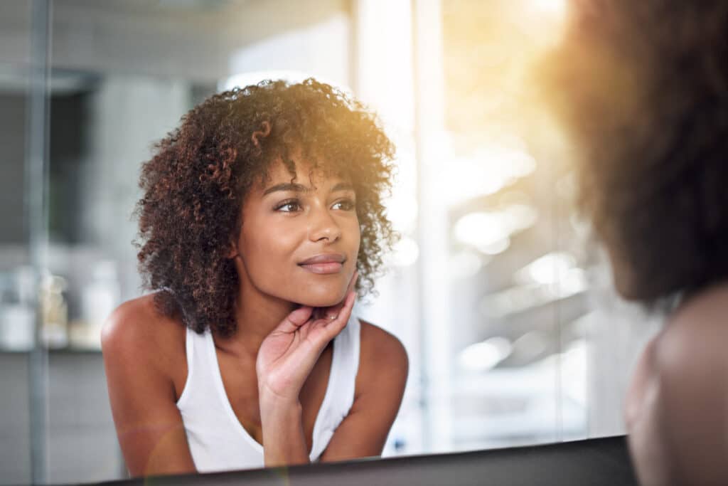 Woman examining her skin in a mirror while considering dermaplaning or microdermabrasion for facial exfoliation.
