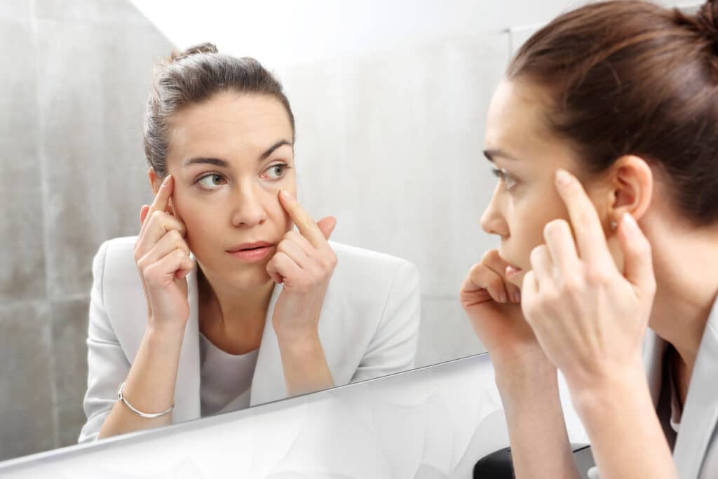 Woman examining the under-eye area in a mirror while considering non-surgical treatments for tired-looking eyes.